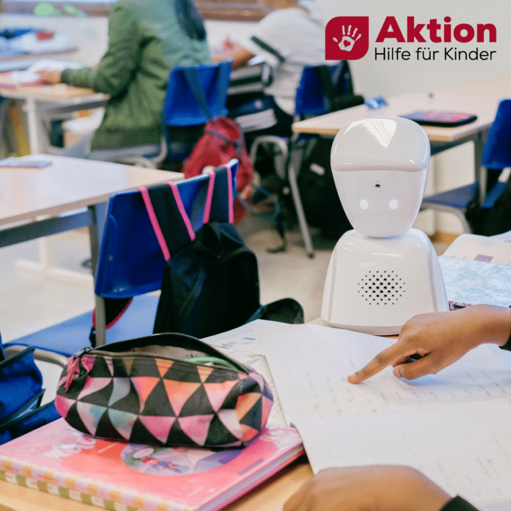 Der Lernroboter sitzt am Tisch und lässt sich von ein/e Schüler*in eine Aufgabe erklären.