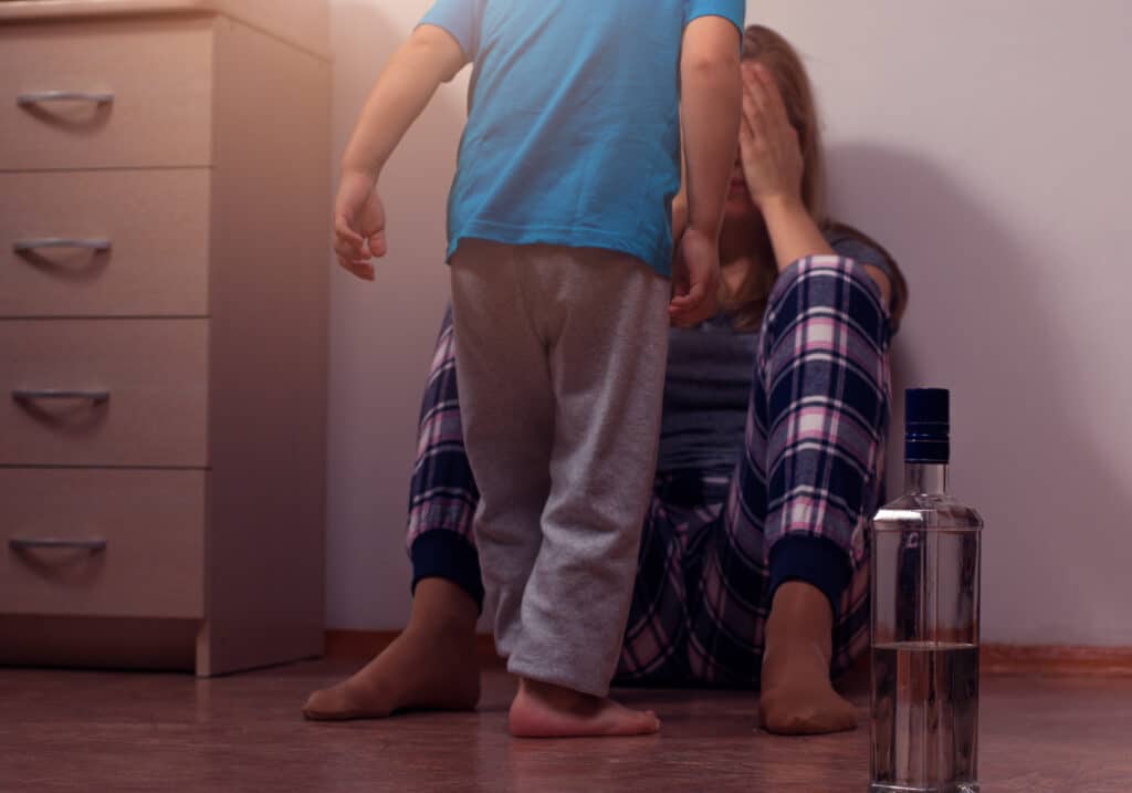 Kind steht vor einer sitzenden Frau, die mit ihren Händen ihr Gesicht verdeckt. neben ihr steht eine Glasflasche mit einer klaren Flüssigkeit.